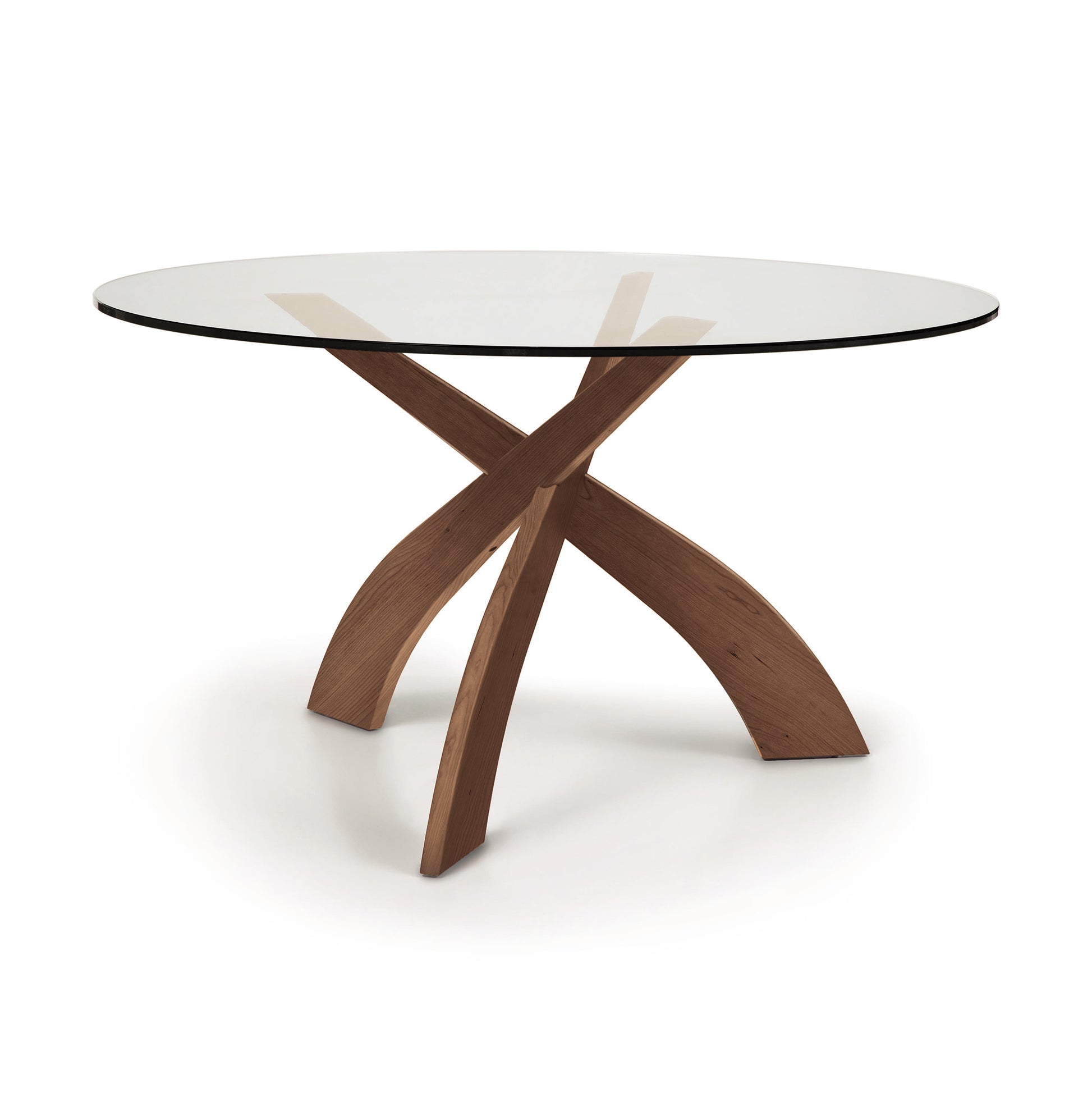 Entwine Round Glass Top Dining Table by Copeland Furniture with intersecting cherry wood legs and a clear tempered glass top, showcasing elegant, modern craftsmanship.