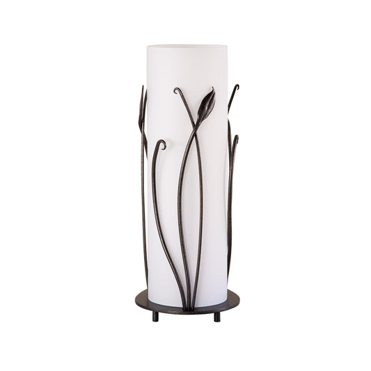 Tall cylindrical Hubbardton Forge Table Lamp with forged leaves, featuring a bronze frame and a round base with three legs.