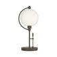 Pluto Table Lamp by Hubbardton Forge featuring a round white glass shade atop a dark metal base and frame, showcasing elegant, modern craftsmanship.