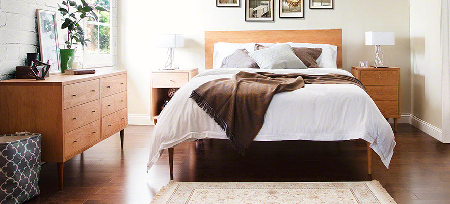 Wooden bed with white bedding and a brown throw, flanked by two nightstands and lamps, accompanied by a dresser on a wooden floor.