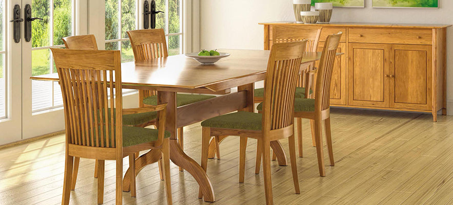 A dining room features a wooden table with six matching wooden chairs. The chairs have green cushioned seats. A sideboard with two drawers and two cabinets stands against the wall. A bowl of green apples sits atop the table, and there are two paintings on the wall.