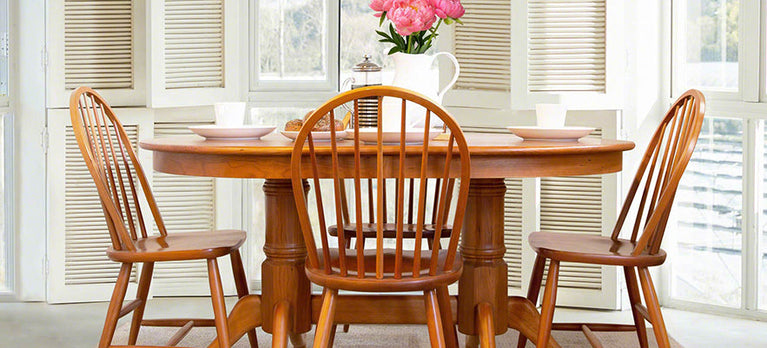 A wooden dining table with three matching chairs is set in front of a white window with shutters. On the table, there is a white pitcher filled with pink flowers, two white cups with saucers, and a metal pot. There is natural light coming in from the windows.
