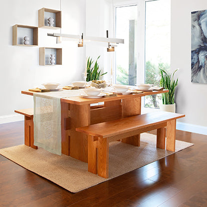 A modern dining room features a wooden table with matching benches. The table is set with white dishes and a beige table runner. Potted plants are placed in the corners. A geometric wall shelf holds small decorative items, and a large abstract painting hangs on the wall.