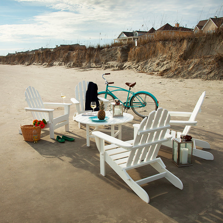 White Adirondack chairs are arranged in a circle on a sandy beach with a round table in the center. Nearby, there's a teal bicycle, a picnic basket, a lantern, and scattered shoes. Houses and dunes are visible in the background under a partly cloudy sky.